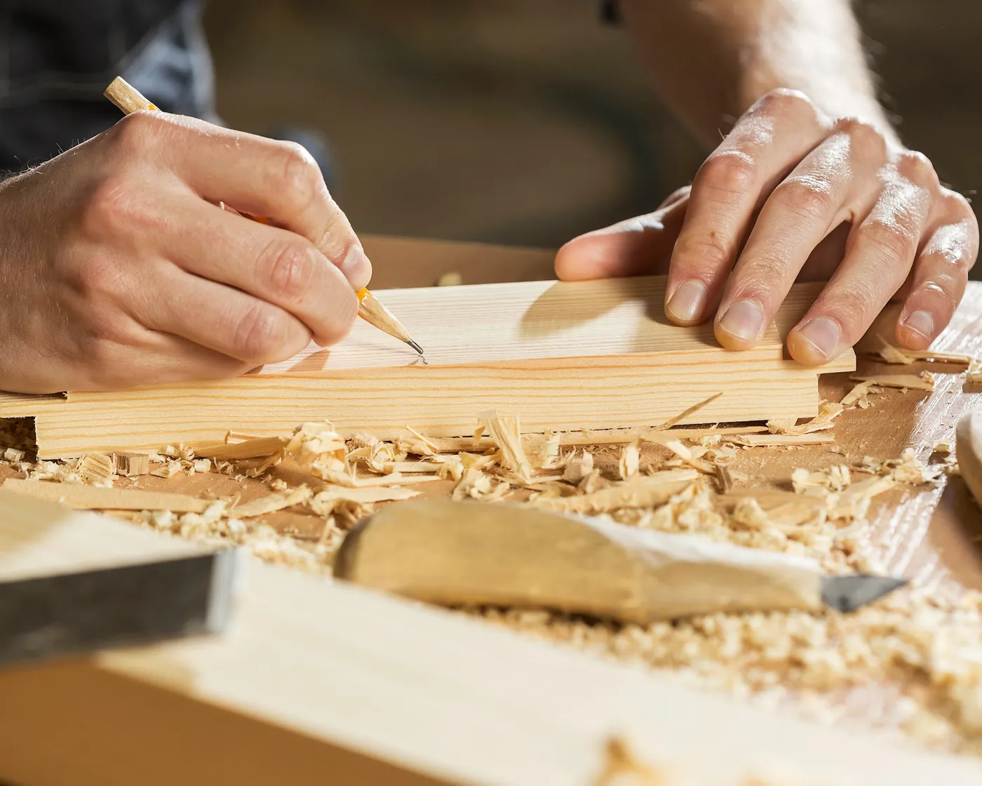 Die Hände eines Tischlers zeichnen mit dem Bleistift ein Stück Holz an, zwischen Hobelspänen.